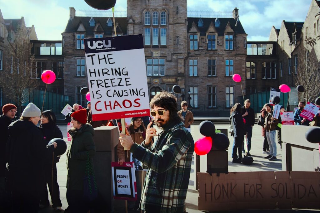 University Staff Strike Over Cost of Living Pay Gap Protesters hold signs outside a building during a demonstration.