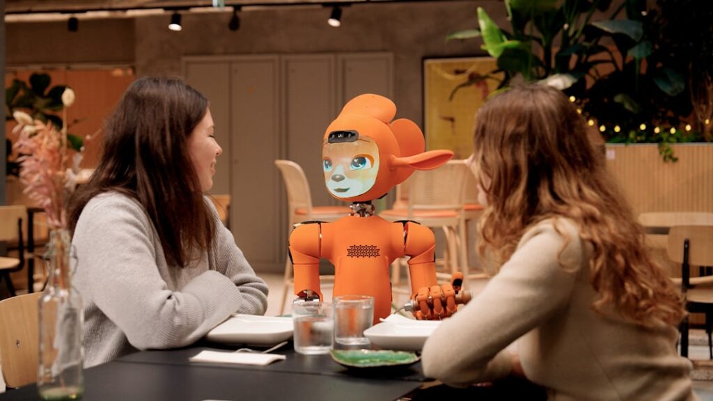 Sierra Acquires Fragment to Expand AI Customer Service Two women talk to an orange robot at a table.