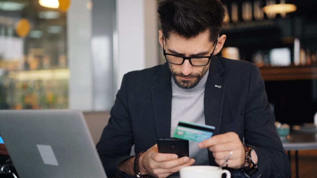Intuit's FedNow Certification Revolutionizes Business Payments a man sitting at a table looking at his cell phone and holding a credit card