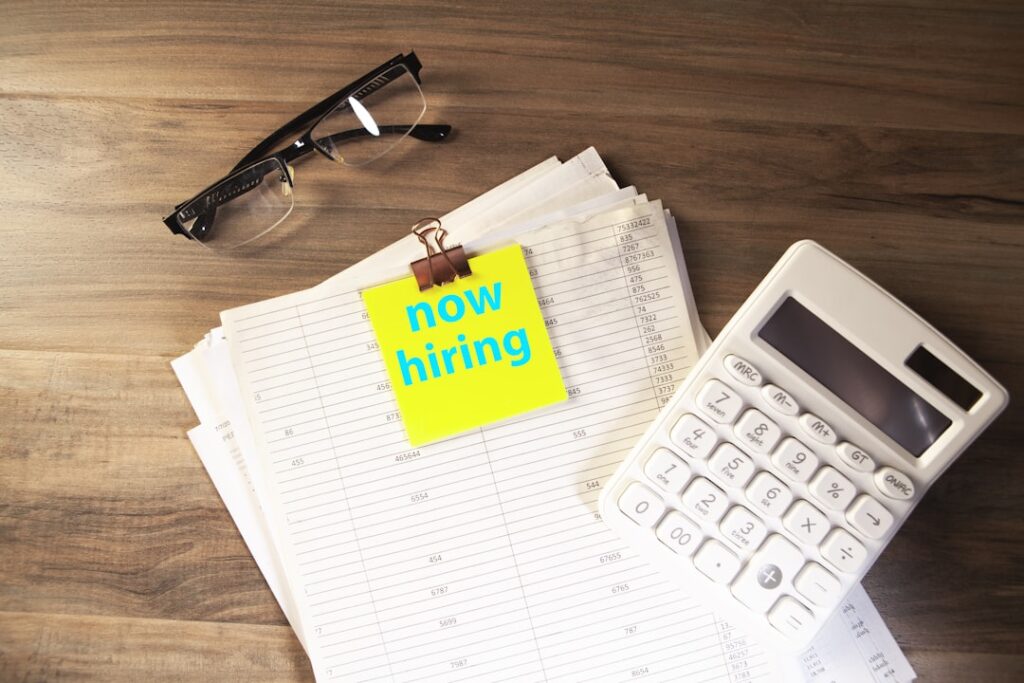 Recruitment Selection Process: Build Your Ideal Team Calculator, glasses, and papers with 'now hiring' note.