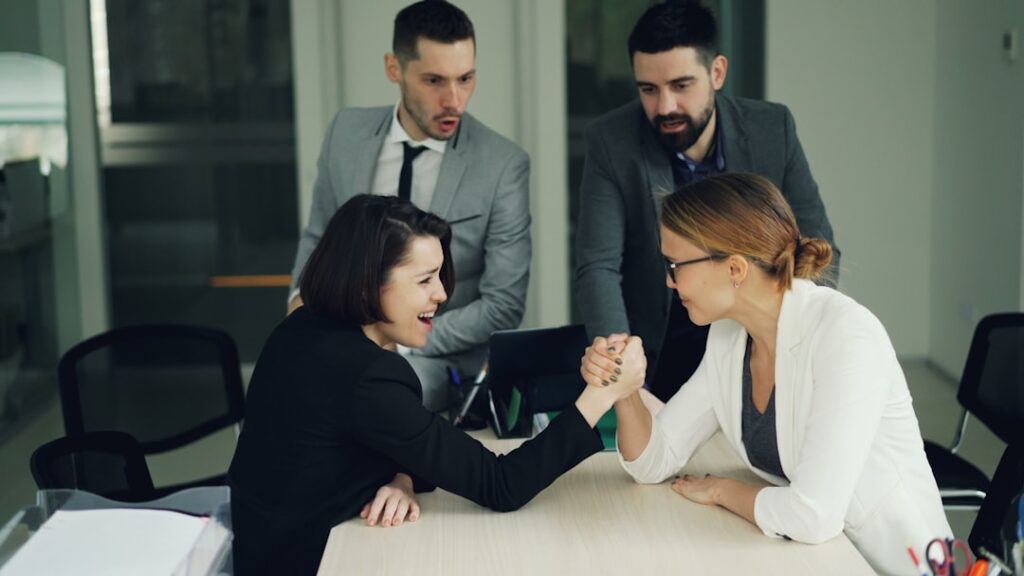 Conflict Management Strategies That Transform Workplace Disputes Two women arm wrestling with men watching them