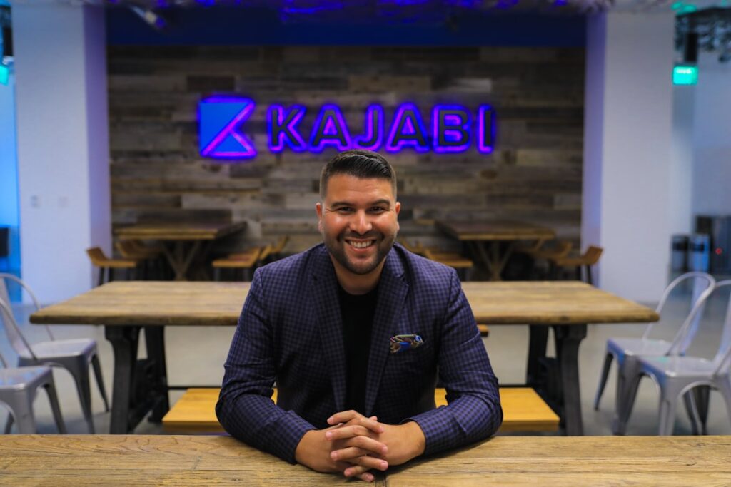 Replit's Masad Defies Acquisition Pressure Amid Cursor Deal man smiling and sitting beside table in front of Kajabi neon signage turned on